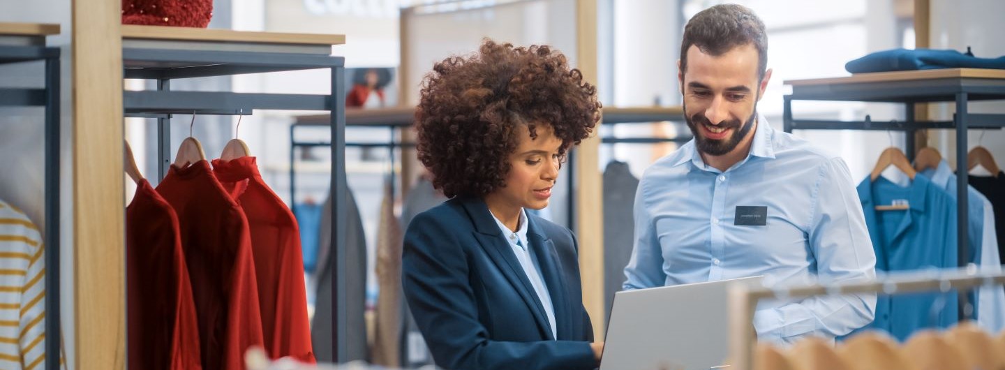 Fashion Management Studium Fashion Managerin erklärt Angestelltem etwas am Laptop zwischen Kleidung