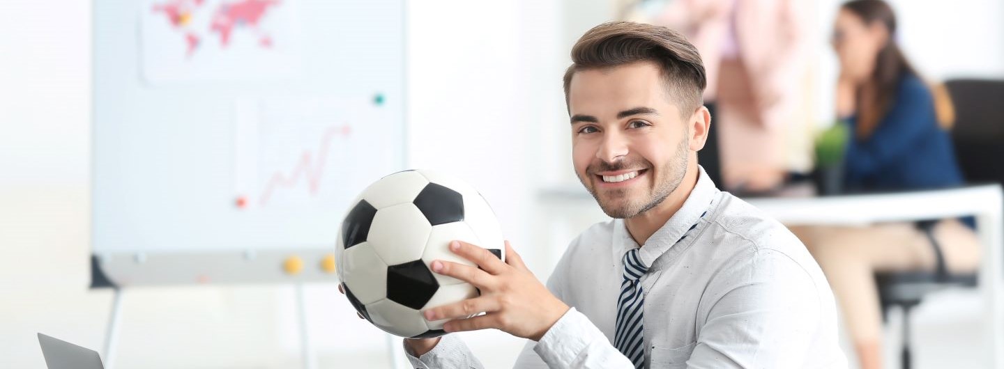 Junger Mann sitzt lächelnd mit einem Fußball in den Händen am Schreibtisch