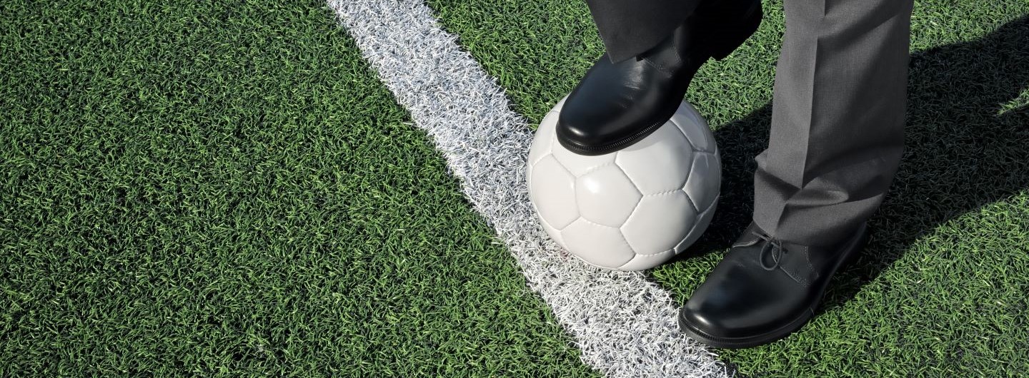 Mann im Anzug steht mit einem Fuß auf Fußball an der Seitenlinie des Feldes