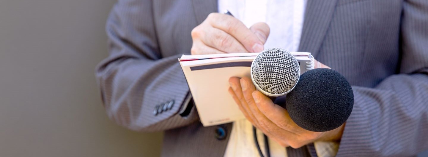 Journalist hält zwei Mikrofone, Zettel und Stift in den Händen
